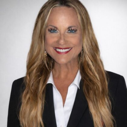 Smiling woman with long blonde hair wearing a black blazer and white shirt, posed in front of a plain light background.