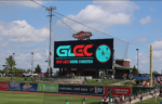 A large digital sign at a baseball stadium displays “Great Lakes Gaming Convention” with people gathered nearby under a partly cloudy sky.