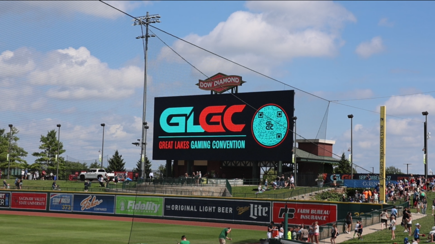 A large digital sign at a baseball stadium displays “Great Lakes Gaming Convention” with people gathered nearby under a partly cloudy sky.