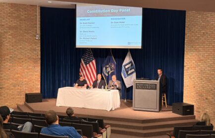 Four panelists sit at a table on stage with U.S. and Michigan flags, as a moderator stands at a podium during Northwood University’s Constitution Day Panel, engaging in thoughtful discussion about America’s Governing Framework.