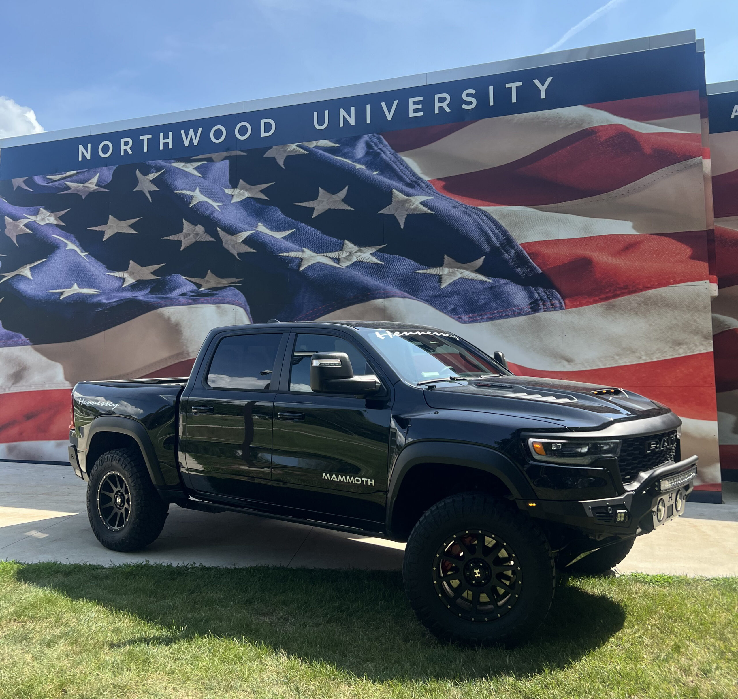 Black pickup truck parked on grass in front of a large American flag mural and Northwood University sign, under a sunny sky, highlighting Sponsor Participation at the International Auto Show.