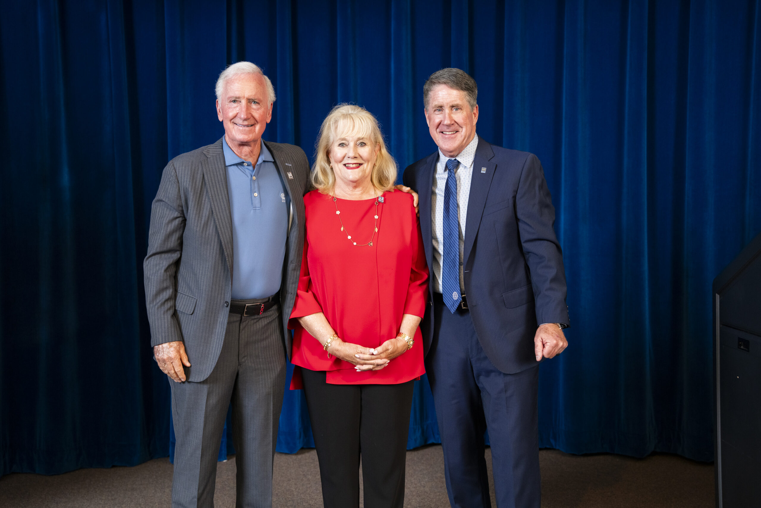 Three adults, two men and one woman, stand side by side in front of a blue curtain. The men wear suits, and the woman wears a red blouse. All are smiling at the camera.