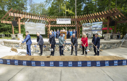 A group of people in business attire use shovels to break ground at an outdoor ceremony, with banners and a pergola in the background.