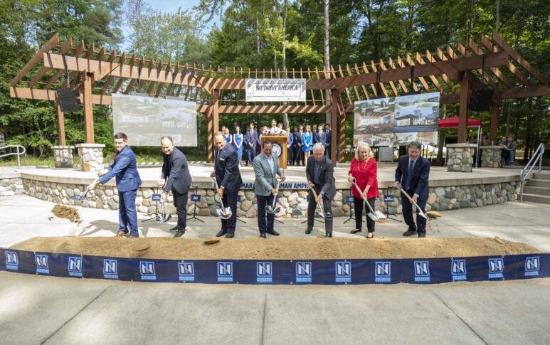 A group of people in business attire use shovels to break ground at an outdoor ceremony, with banners and a pergola in the background.