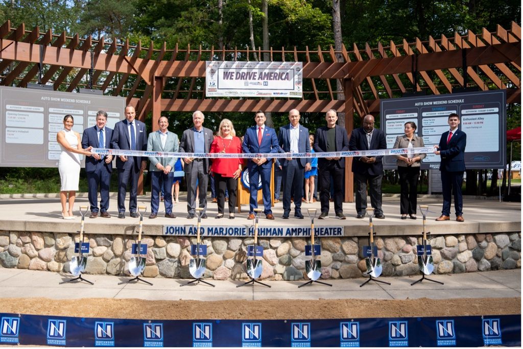 A group of people in formal attire stands behind a ribbon at an outdoor amphitheater stage for a ribbon-cutting ceremony, with banners and schedules displayed in the background.
