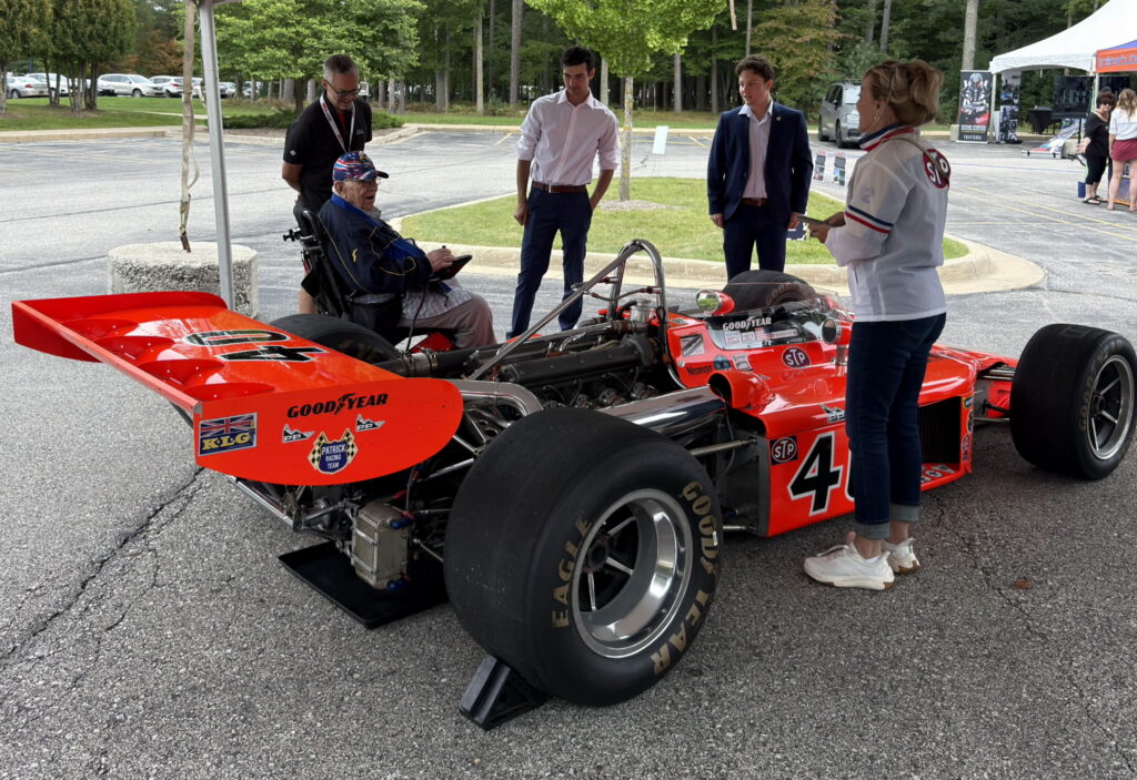 A man in a wheelchair and three others stand around The Honker, a bright orange vintage race car numbered 4, in an outdoor parking lot at the Northwood Auto Show.