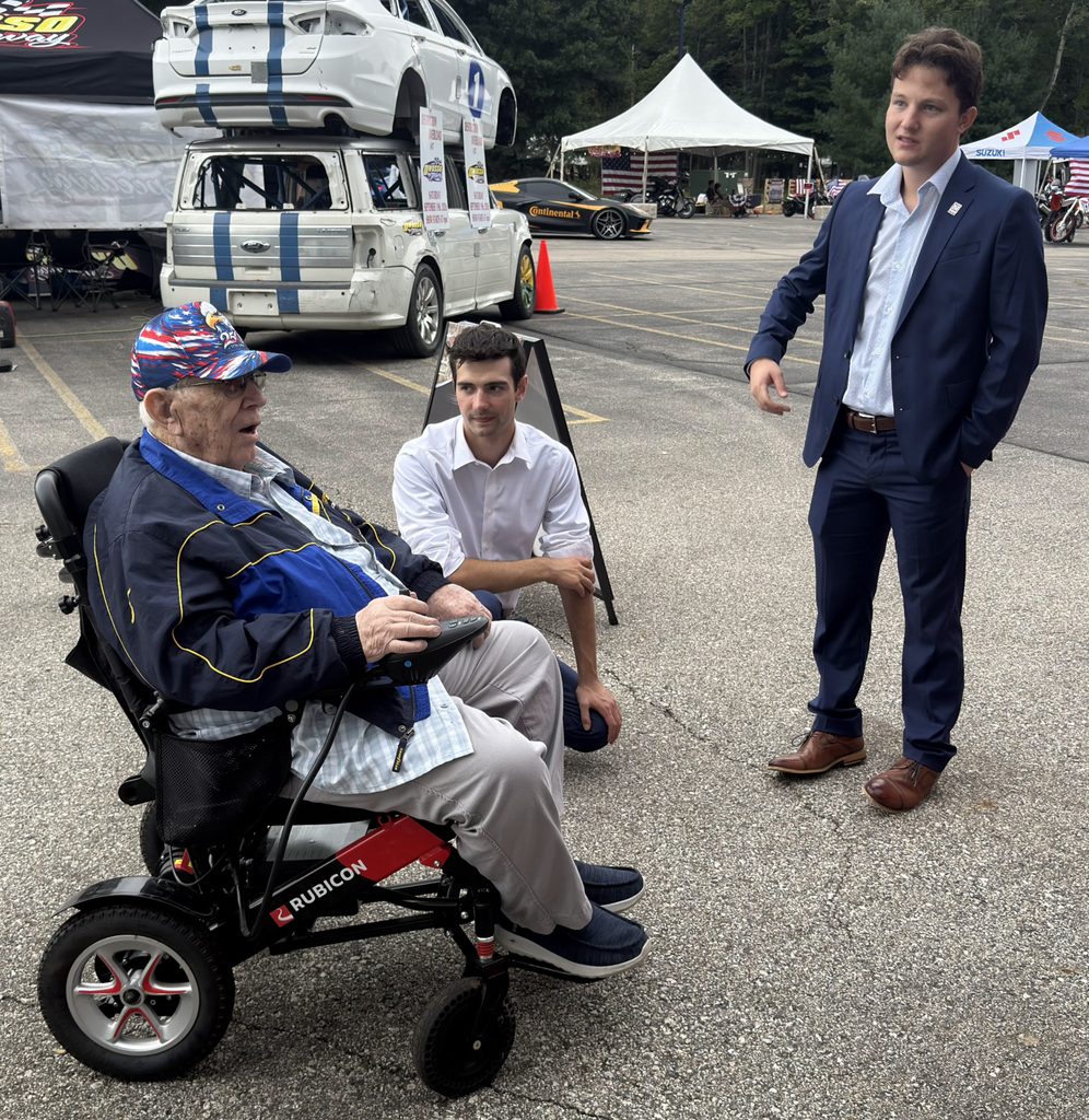 An older man in a wheelchair speaks with two younger men standing and kneeling in a parking lot near several vehicles.