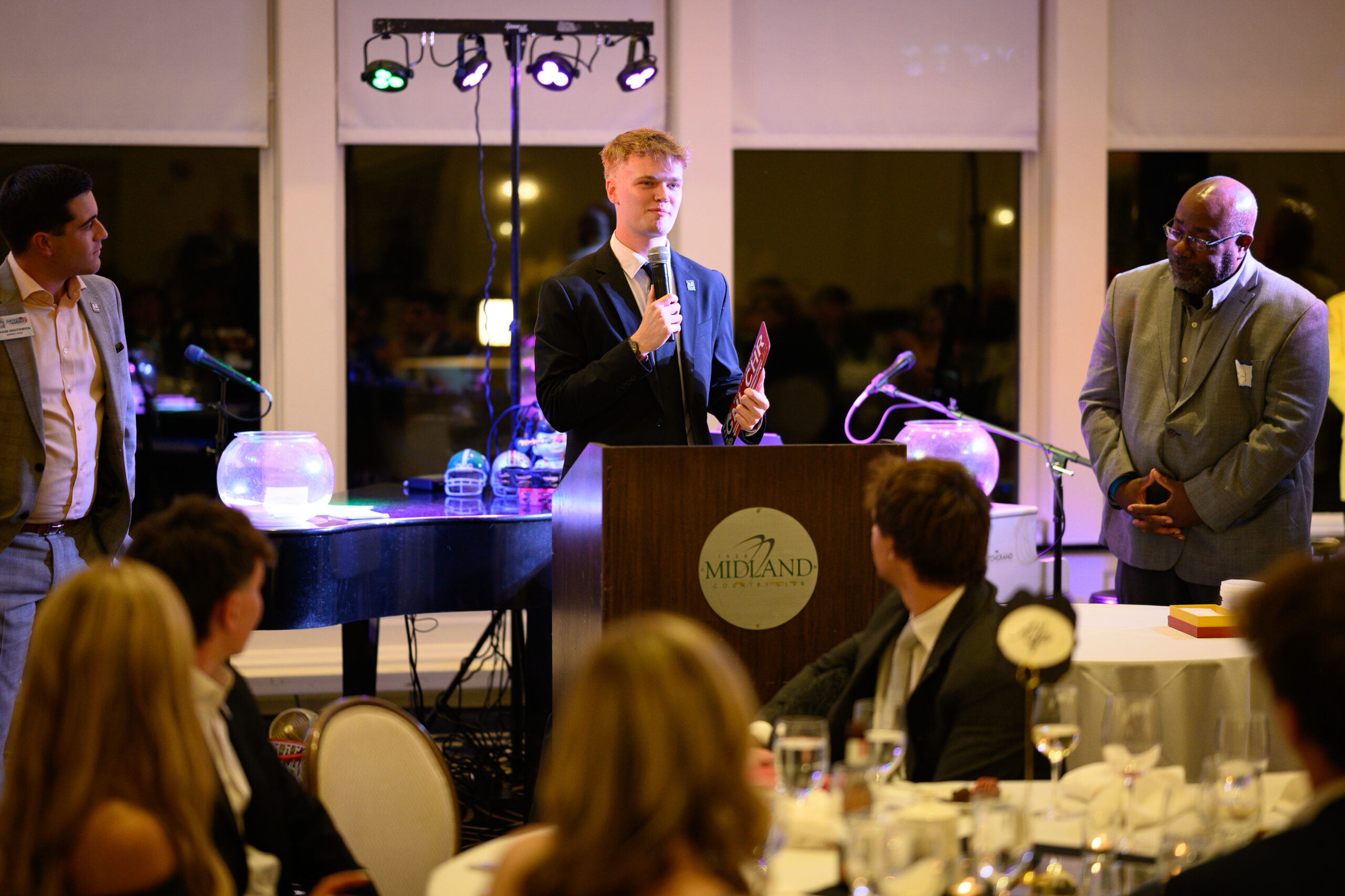 Nick Porter, the 2027 General Chair, speaks into a microphone at a Midland-branded podium, flanked by two men during an auto show event with seated guests.