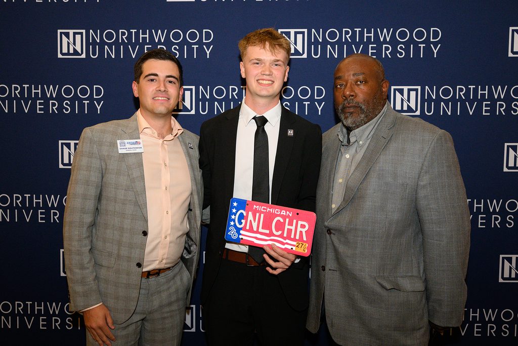 Three students, including Nick Porter, stand in front of a Northwood University backdrop, with the man in the center holding a Michigan license plate that reads “GNLCHR”.
