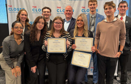 A group of students and two mentors pose in front of a branded “The Closer” backdrop, with two students holding “Sales Role Play” award certificates.