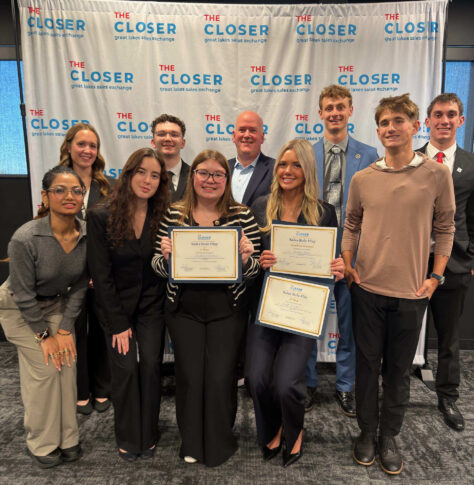 A group of students and two mentors pose in front of a branded “The Closer” backdrop, with two students holding “Sales Role Play” award certificates.