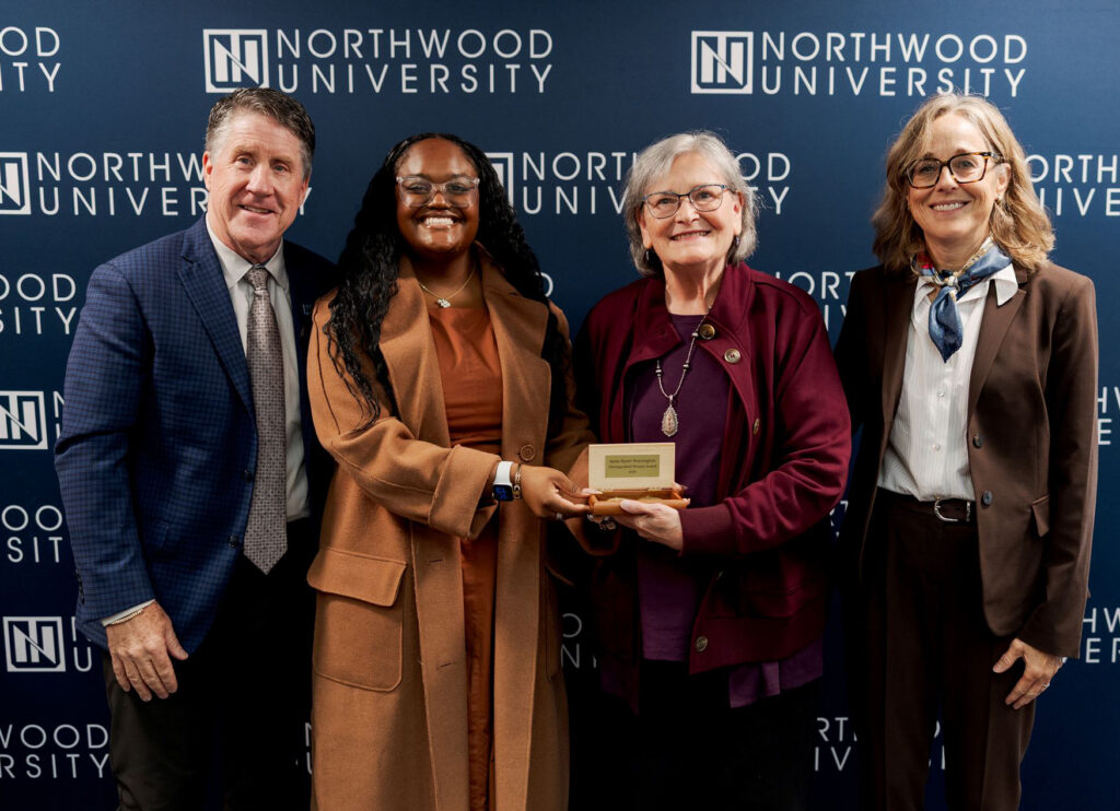 Four people stand before a Northwood University backdrop as one person presents a small award plaque to another during a women’s initiative event.
