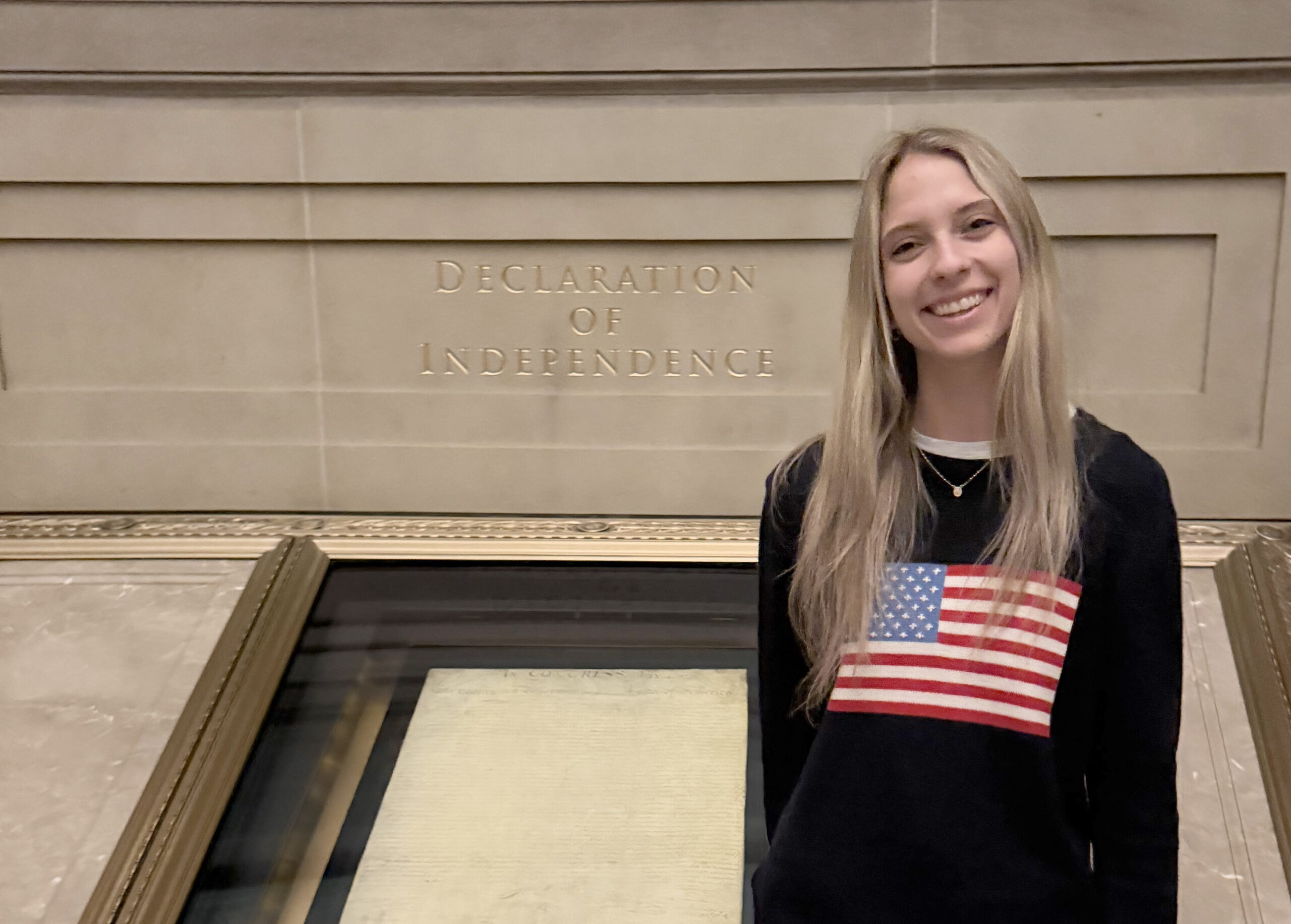 A smiling woman in an American flag sweater stands before the Declaration of Independence at the National Archives, capturing footage for a video contest.
