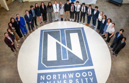 A large group of students in professional attire standing in a circle around the Northwood University logo on a floor.