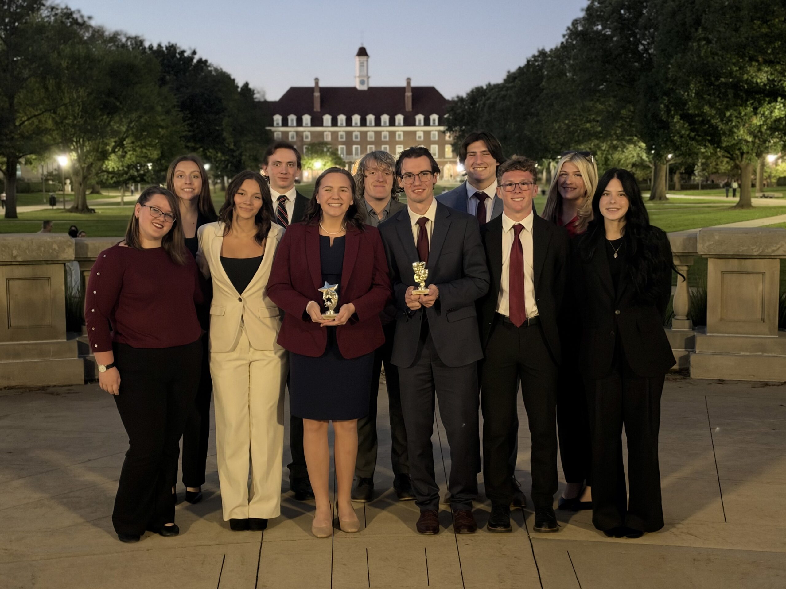 A group of students in professional attire posing with two small trophies in front of a university building at dusk.