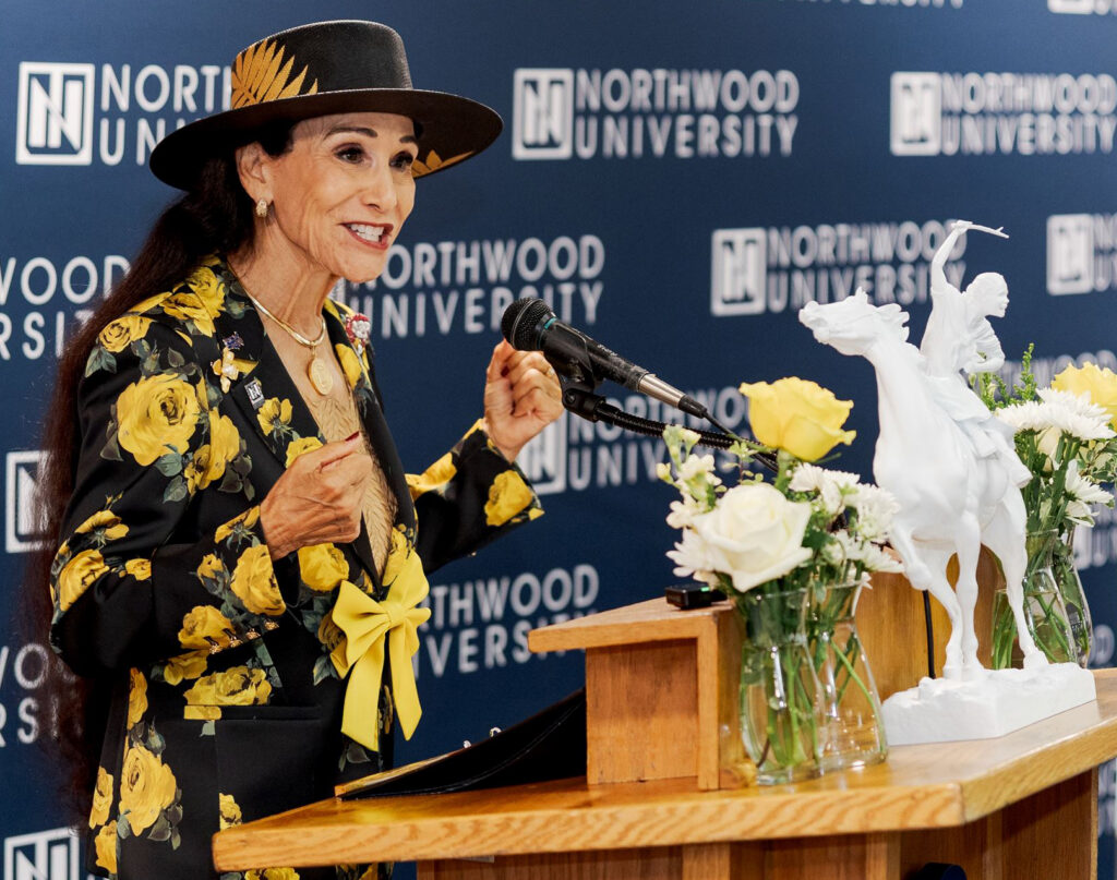 A woman in a black and yellow floral blazer and hat speaks at a wooden podium at a Northwood University women’s initiative, with a white equestrian statue and flower vases nearby.