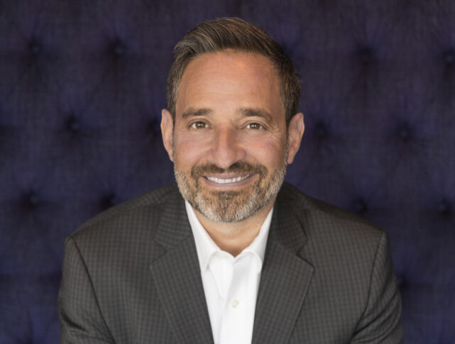 A middle-aged man with a beard, wearing a white shirt and dark blazer, smiling at the camera.