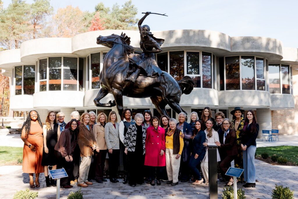 A group of women posing in front of a bronze statue symbolizing women’s courage on a rearing horse at the Northwood University campus.