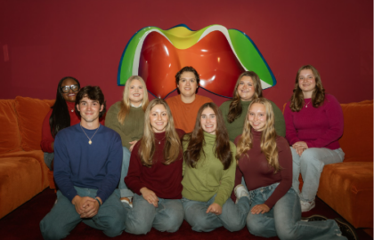 A group of nine young adults posing together in front of a colorful wall sculpture and orange seating.