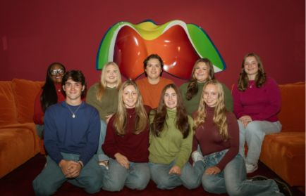 A group of nine young adults posing together in front of a colorful wall sculpture and orange seating.
