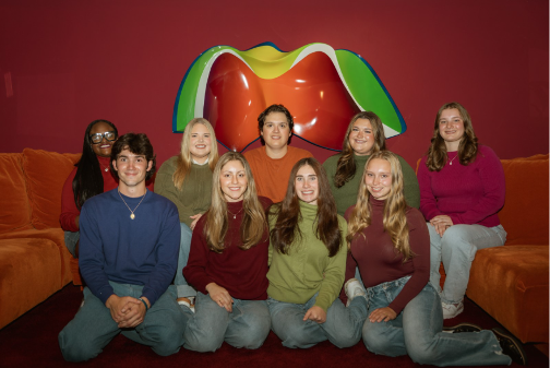 A group of nine young adults posing together in front of a colorful wall sculpture and orange seating.