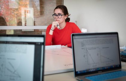 A woman in glasses sits at a table with laptops displaying charts and papers in front of her, exuding the thoughtful demeanor of a Master of Business Administration.