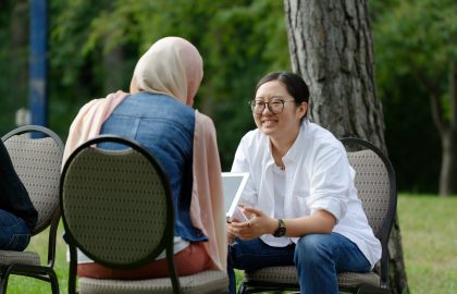 Two people sitting in outdoor chairs, one wearing a hijab and denim jacket, the other in glasses and a white shirt, engaging in conversation near tree.