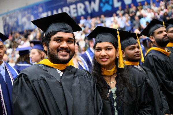 Graduates in caps and gowns smile at a commencement ceremony with a crowd and banner in the background.