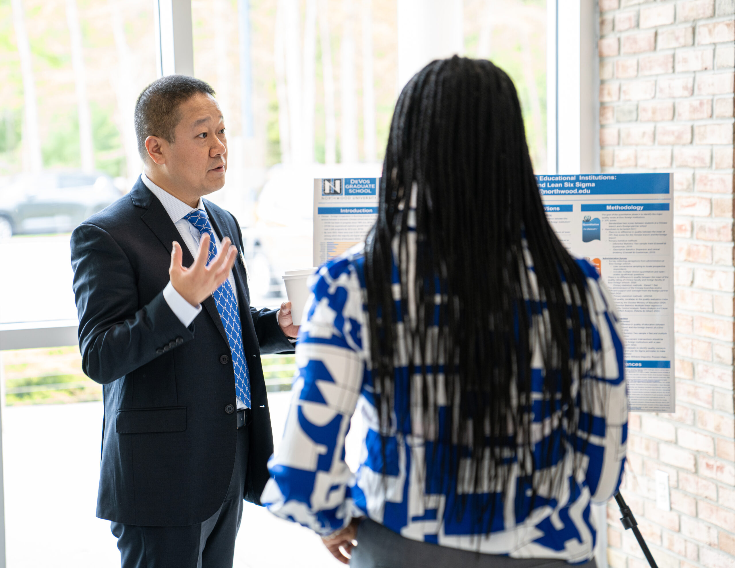 Two people stand in front of a presentation board. The man gestures while holding a coffee cup; the woman, with braided hair, listens. They are in a brightly lit room.