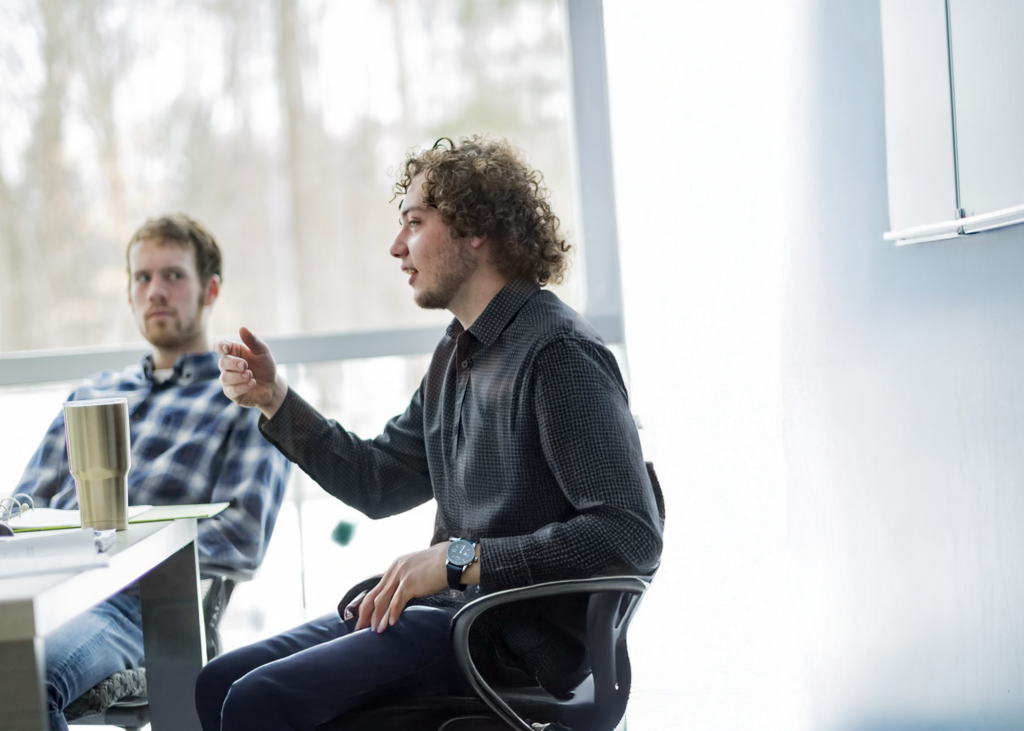 In an office setting, one individual gestures passionately while speaking, the other listens attentively. A window bathes the room in light, highlighting a table adorned with a tumbler and notebook.