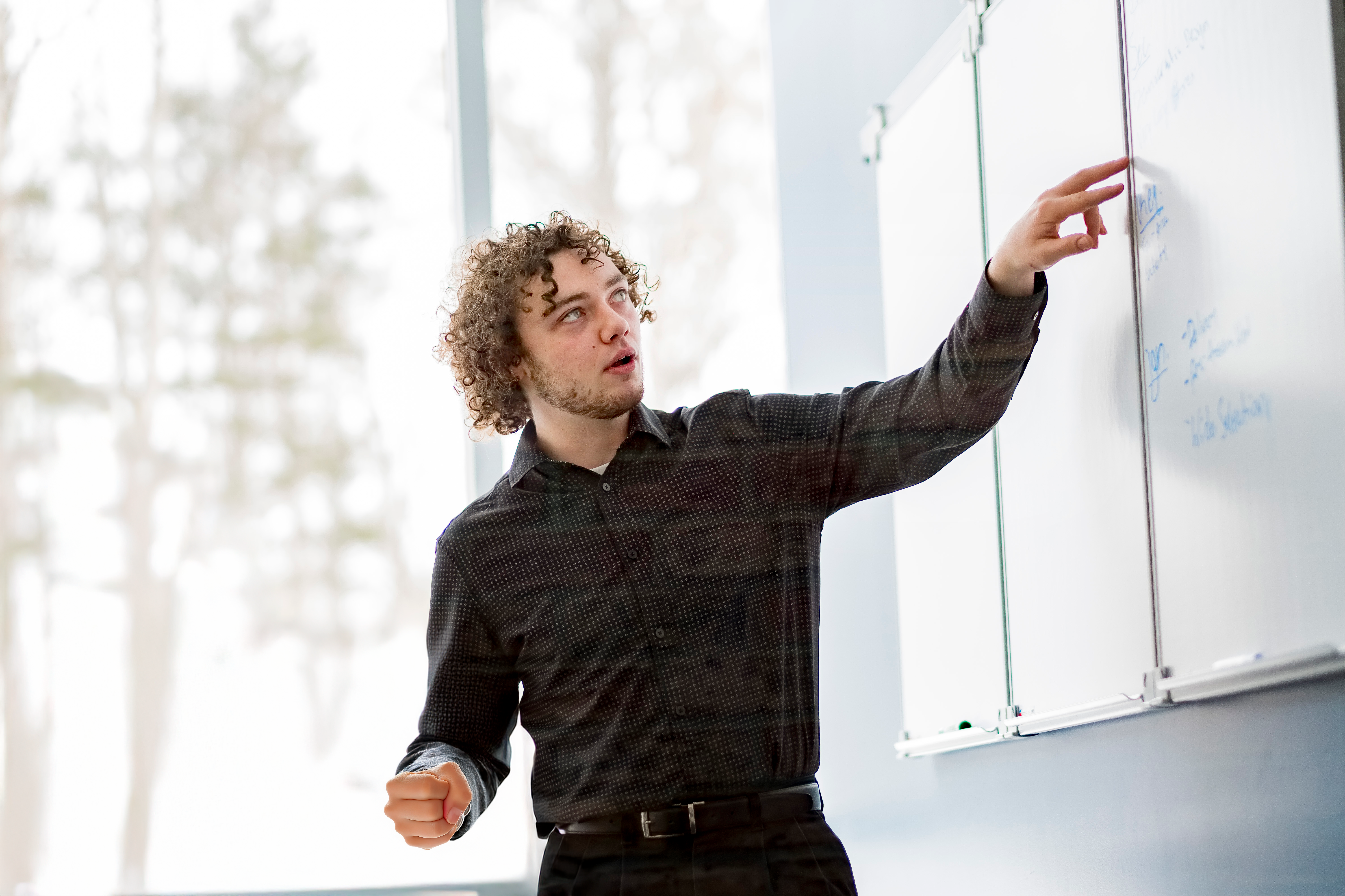 A person with curly hair points at a whiteboard in an office setting, speaking to an unseen audience.