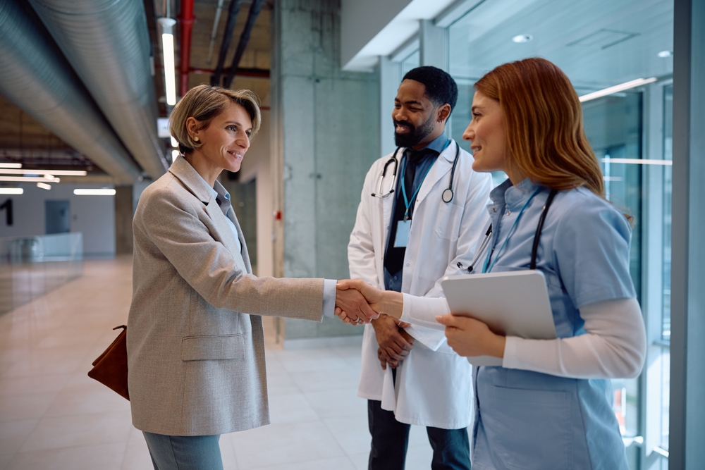 A woman in business attire shakes hands with a nurse while a doctor stands nearby in a hospital hallway.