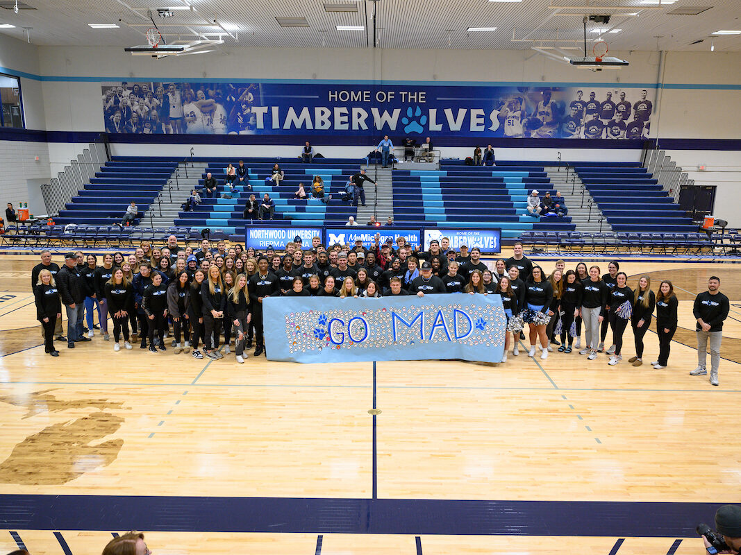 A group photo of numerous Northwood athletes on a basketball court holding a "GO MAD" blue sign with blue bleachers spelling "NU" behind them.