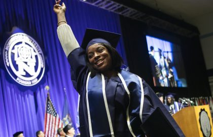 Graduate in cap and gown raises arm in celebration on stage at commencement ceremony, surrounded by blue curtains and flags, as international partners look on with pride.