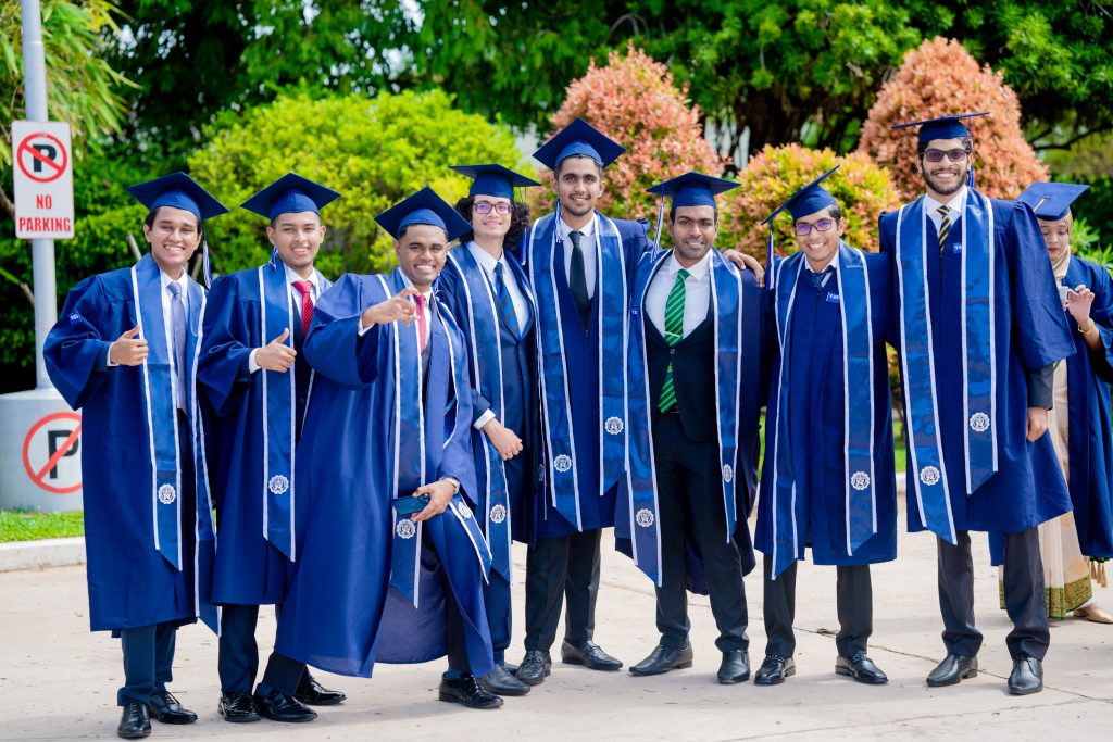 A group of eight graduates in blue gowns and caps pose outdoors, some smiling and giving thumbs up.