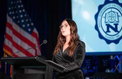 A woman in a black outfit speaks at a podium with a microphone. An American flag and a university logo are in the background.