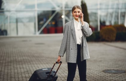 A person in a gray blazer is talking on the phone while pulling a suitcase outdoors.