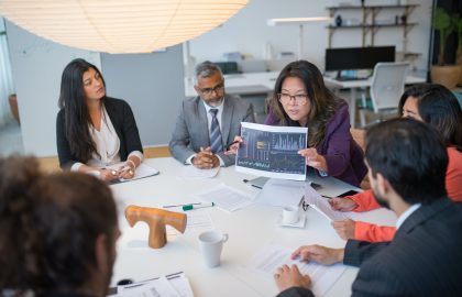 A business meeting with a group of people seated at a table. A woman shows a chart to the group, discussing data. Papers and coffee cups are on the table.