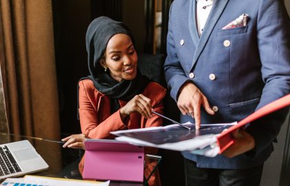 Two people in a business setting reviewing documents at a table with a laptop and tablet present.