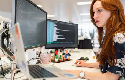 A person with red hair works intently at a desk with multiple computer screens displaying code. The workspace is decorated with small figurines.