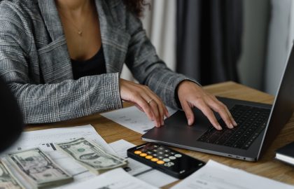 A person in a plaid blazer uses a laptop at a desk with cash, receipts, and a calculator displaying 65.