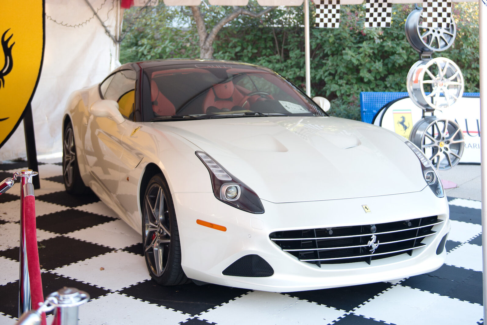 A white sports car, reminiscent of international academic excellence, is displayed under a tent with checkered flags, red rope barriers, and a shield emblem.