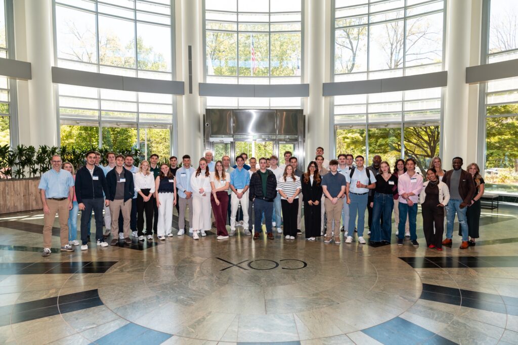 A large group of people stands in a semi-circle in a bright indoor atrium with large windows, marble floors, and the word “COX” on the floor, gathered for the Northwood University Computer Automotive Retail Simulation event.