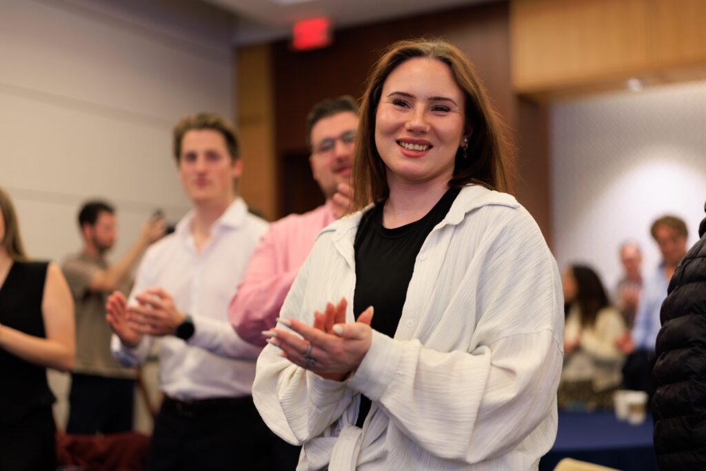 A woman in a white shirt stands and smiles while clapping in a room with other people, possibly celebrating a successful Northwood University Computer Automotive Retail Simulation. The background is slightly blurred.