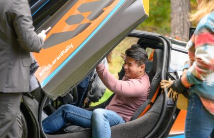 A person sits inside a car with open gullwing doors, smiling, surrounded by others standing nearby. The sleek vehicle is partially visible with #McLarenRally on it, showcasing the brilliance of automotive marketing in action.
