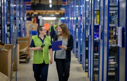 Two people in a warehouse, one holding a clipboard and the other a handheld device, walk and talk between rows of shelves filled with various items.