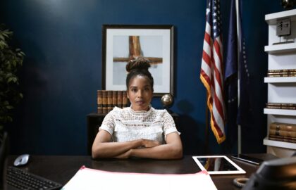 A woman sits at a desk with folded arms, exuding an air of leadership in an office adorned with flags and bookshelves, embodying her dedication to public safety and service.