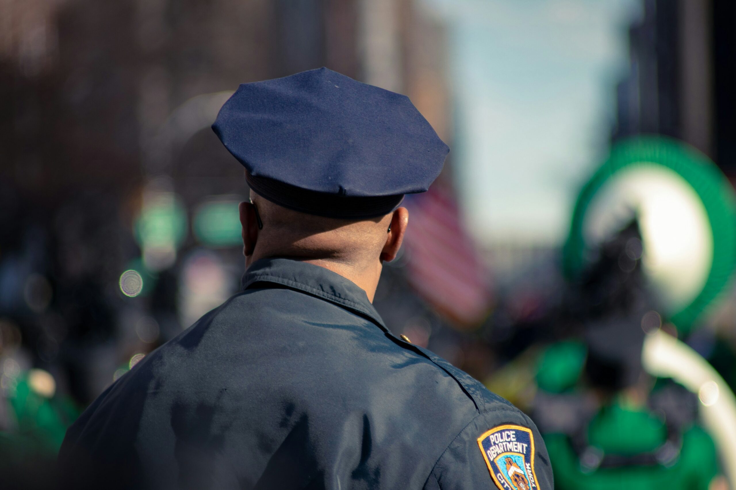 A uniformed police officer stands with his back to the camera, observing a public event with a flag and green decorations in the background.