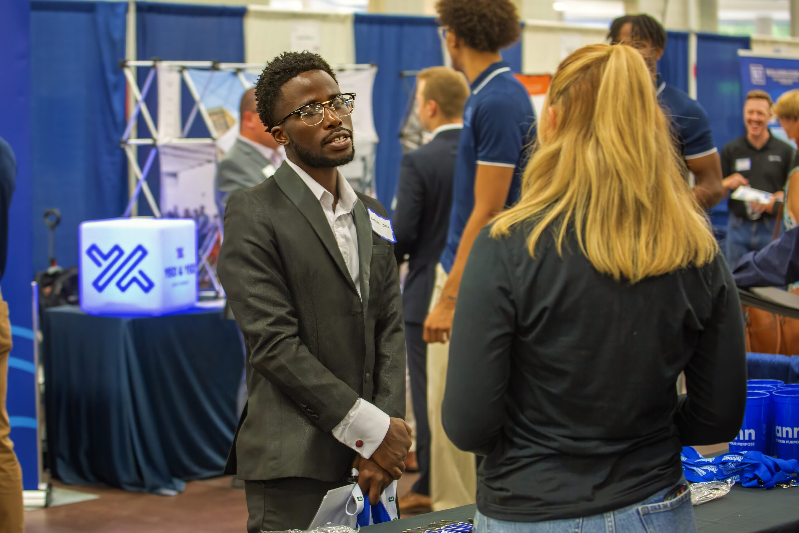 A man in a suit talks to a woman at a busy job fair, with booths and company banners visible in the background.