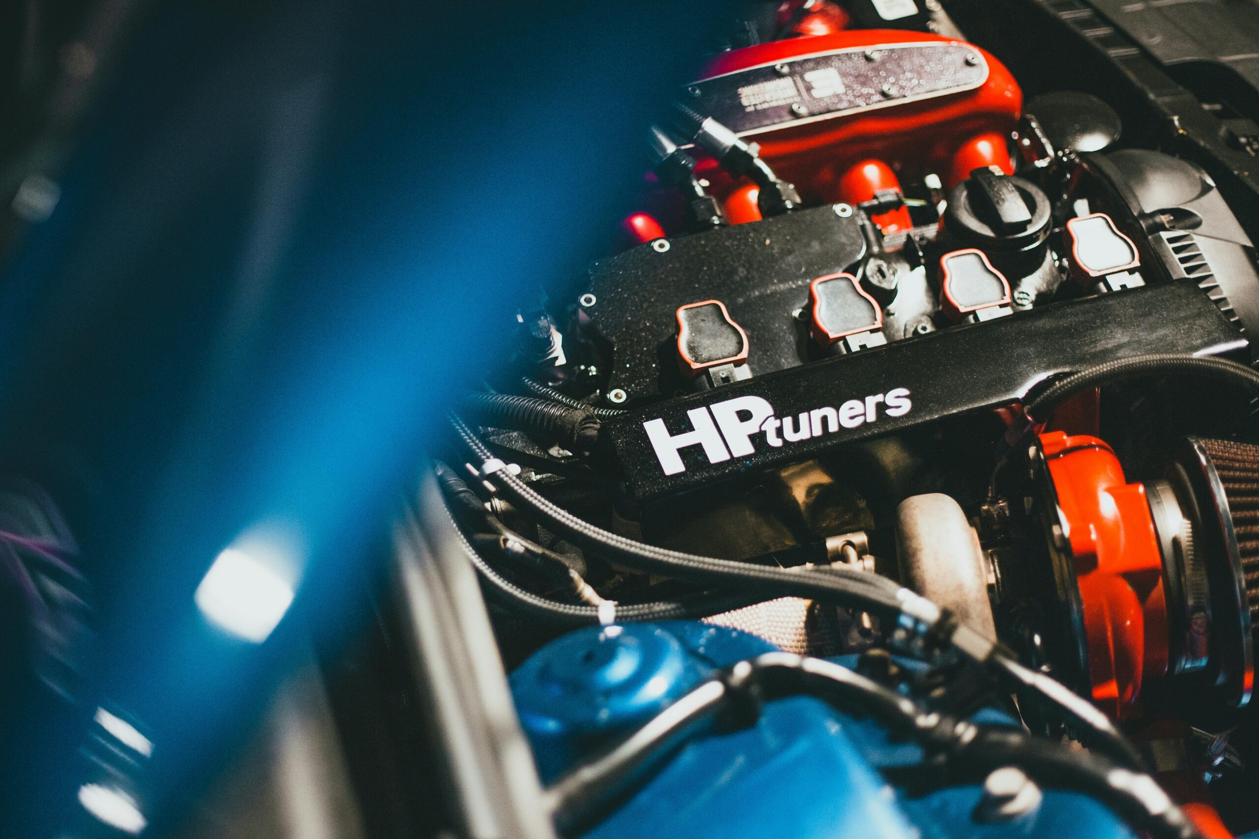 Close-up of a car engine with an HPtuners logo on the cover, showing various components in a well-lit environment.
